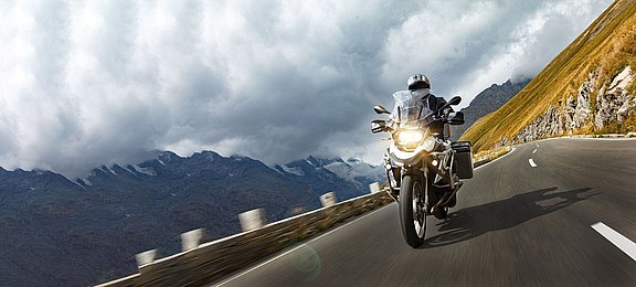 Foto: Ein Motorradfahrer auf einer Reiseenduro fährt mit eingeschaltetem Licht zügig eine Bergstraße entlang.