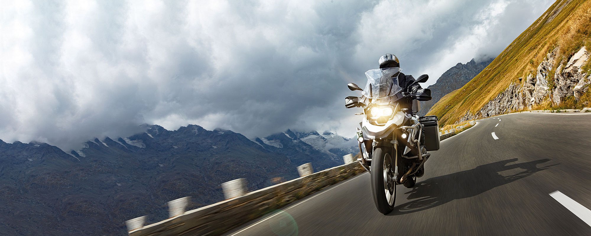 #htmlcaption-3 Foto: Ein Motorradfahrer auf einer Reiseenduro fährt mit eingeschaltetem Licht zügig eine Bergstraße entlang.