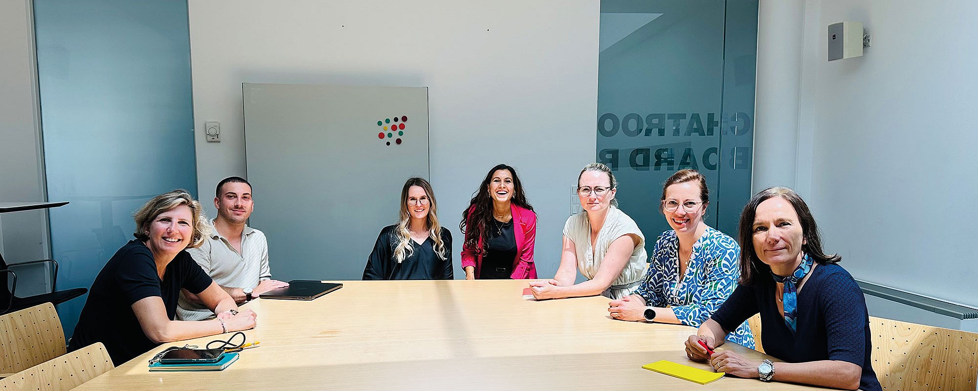 #htmlcaption-14 Foto: Sieben Personen sitzen gemeinsam an einem Besprechungstisch in einem hellen Konferenzraum. Die freundliche Gruppenaufnahme vermittelt Zusammenarbeit, Austausch und Teamgeist in einer professionellen Arbeitsatmosphäre mit moderner Büroeinrichtung.